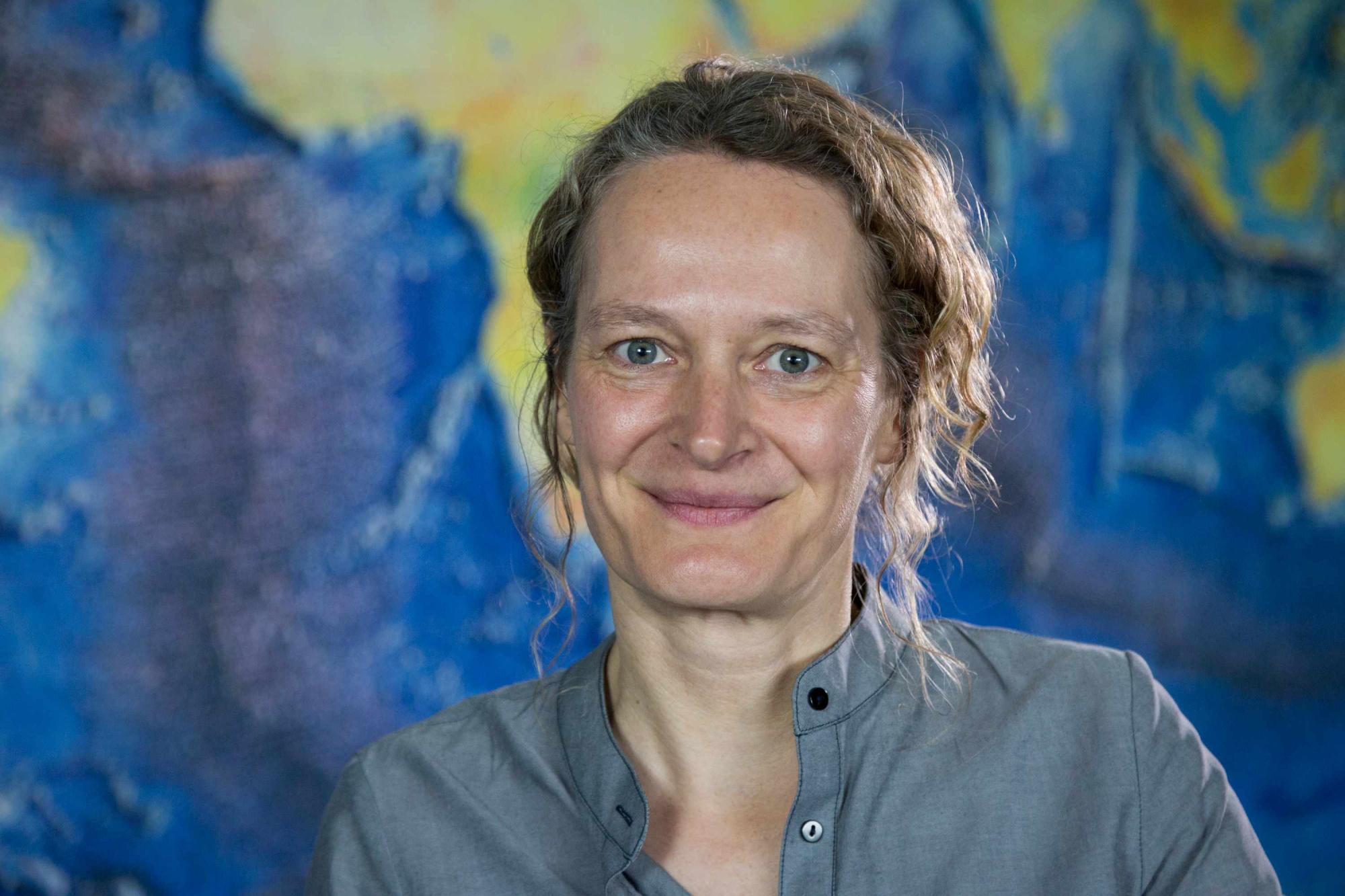 Photograph of Barbel Honisch. Woman smiling at the camera in grey-blue shirt in front of topographic map.