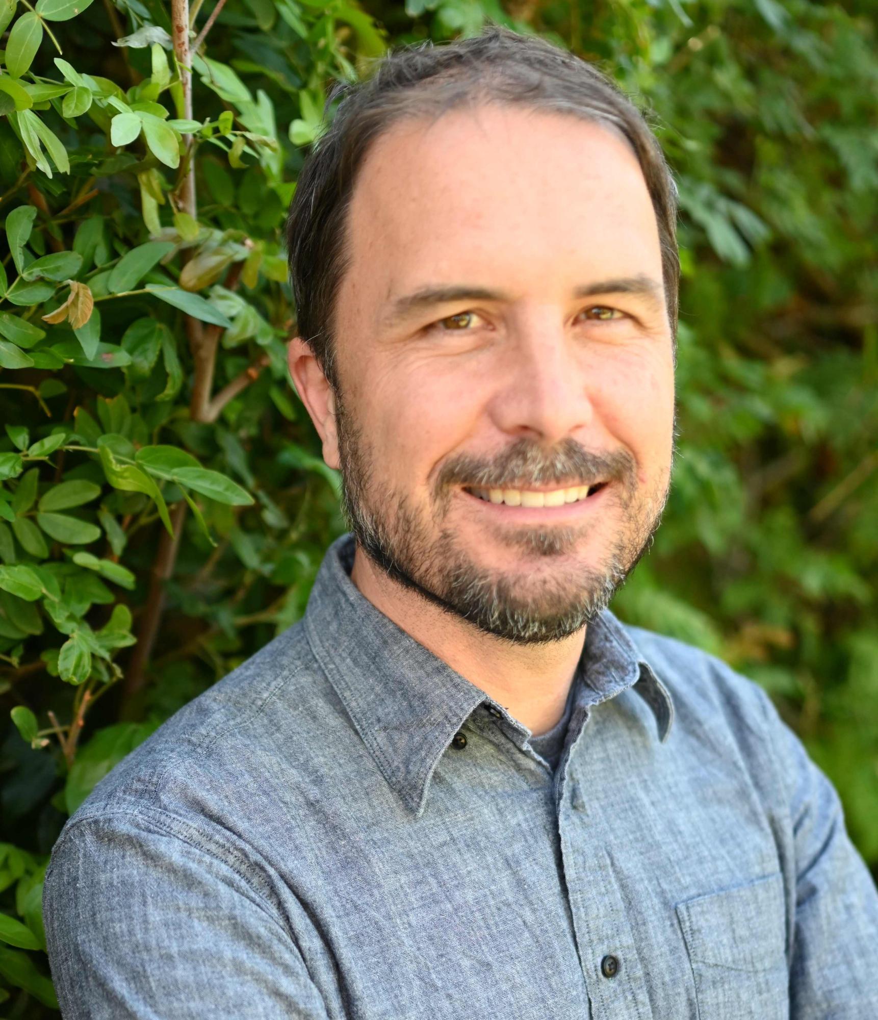 Photograph of Ben Poulter. Man smiling at camera in blue shirt in front of green foliage.