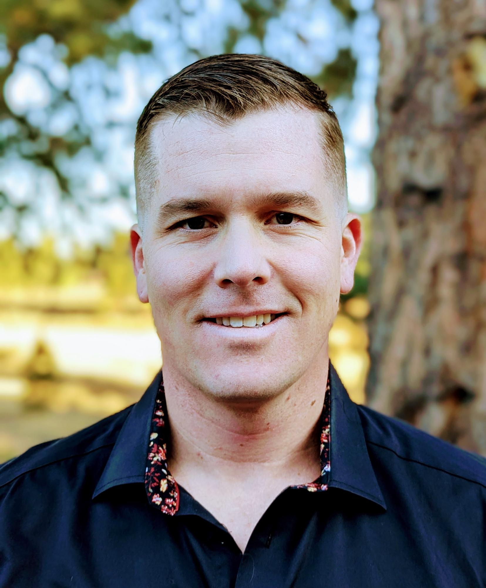 Photograph of J. Blake Clark. Man smiling at camera with navy blue shirt in front of tree in sunshine.