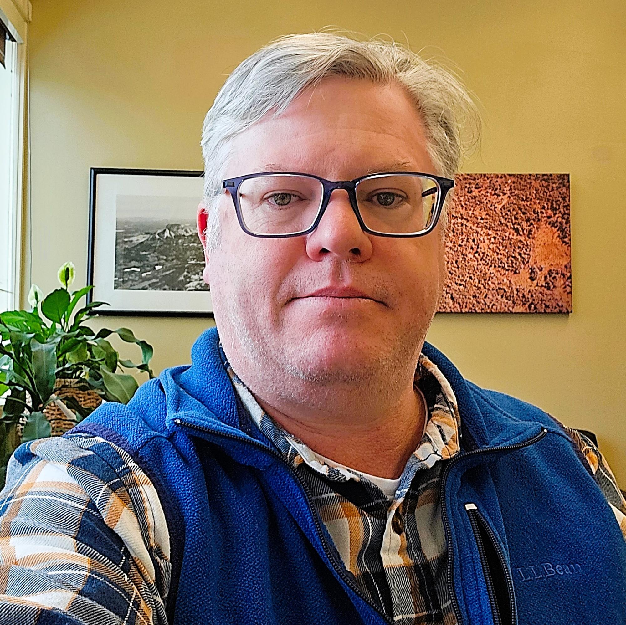 Photograph of Daniel Hayes in front of plant and pictures of landscapes. Man smiling wearing glasses and vest.