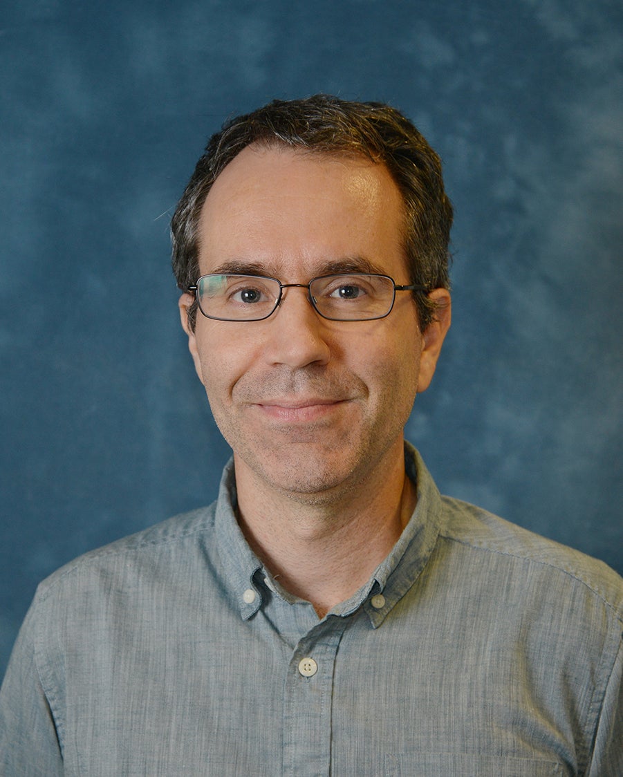Photograph of Dana Royer, man in light blue shirt with blue background, with glasses, smiling.