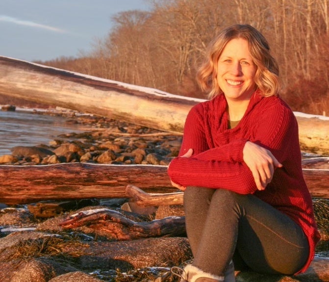 Photograph of Heather Benway. Woman smiling at camera in red sweater in front of snowy beach and wood logs.
