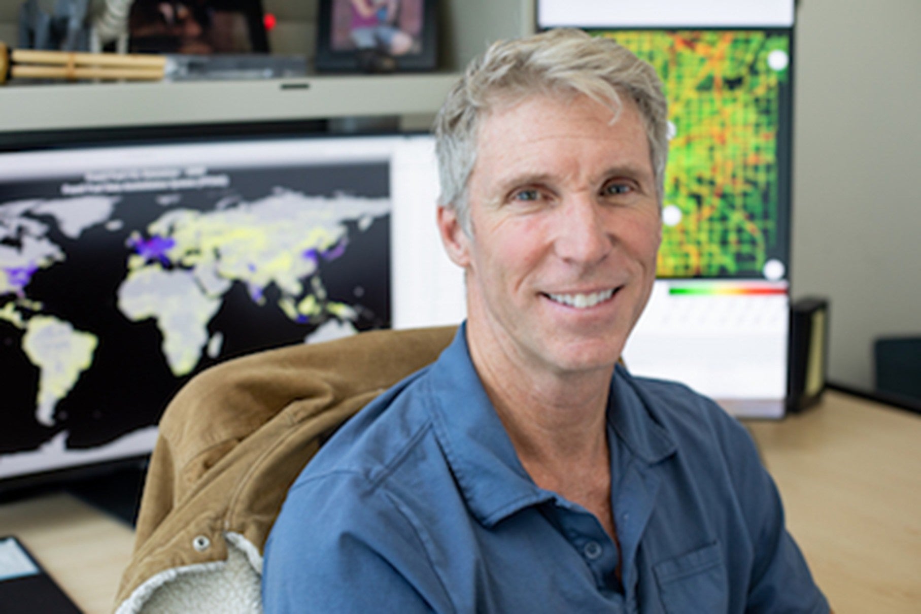 Photograph of Kevin Gurney, man, in front of maps. Man is smiling, seated in chair.